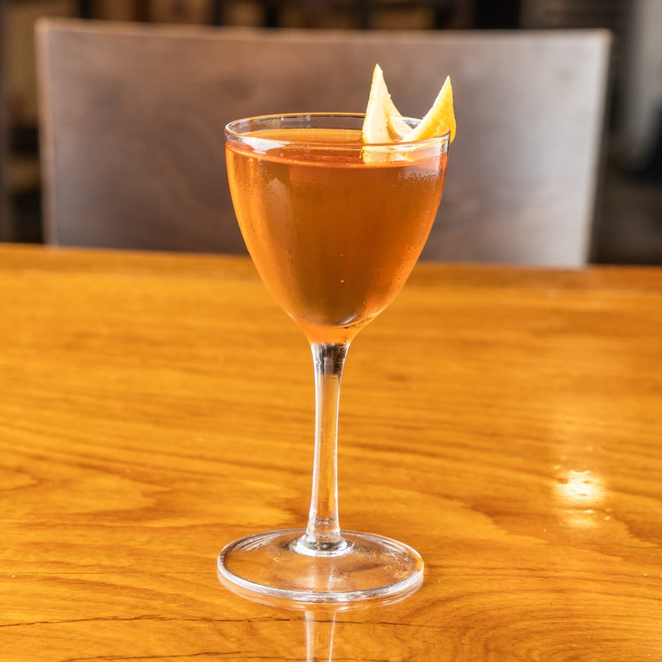 A chilled amber cocktail in a coupe glass garnished with orange peel on a wooden bar.
