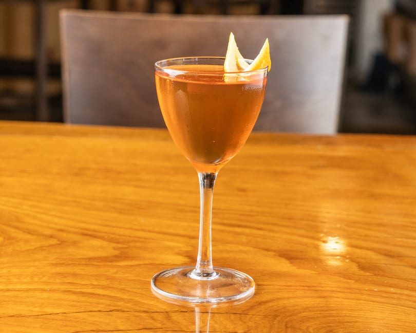 A chilled amber cocktail in a coupe glass garnished with orange peel on a wooden bar.