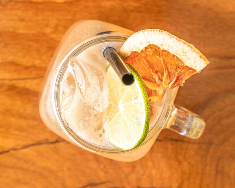 Overhead view of a drink with ice, lime, and dried orange in a glass jar on wood.