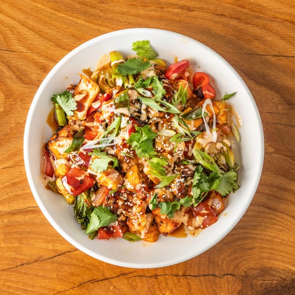 Vibrant chicken and vegetable bowl with sauces, cilantro, and sesame seeds on a wooden table.