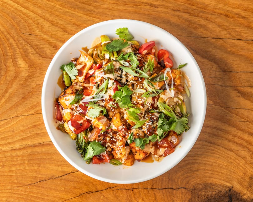Vibrant chicken and vegetable bowl with sauces, cilantro, and sesame seeds on a wooden table.