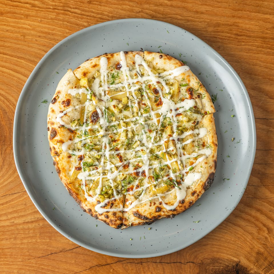 Round flatbread pizza topped with potato slices, dill, and white sauce on a grey plate.