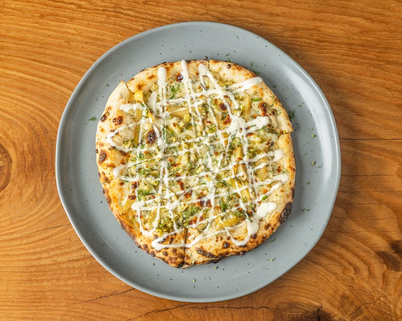 Round flatbread pizza topped with potato slices, dill, and white sauce on a grey plate.