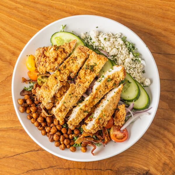 Crispy fried chicken salad with roasted chickpeas, cucumber, tomatoes, and crumbled cheese on a white plate.