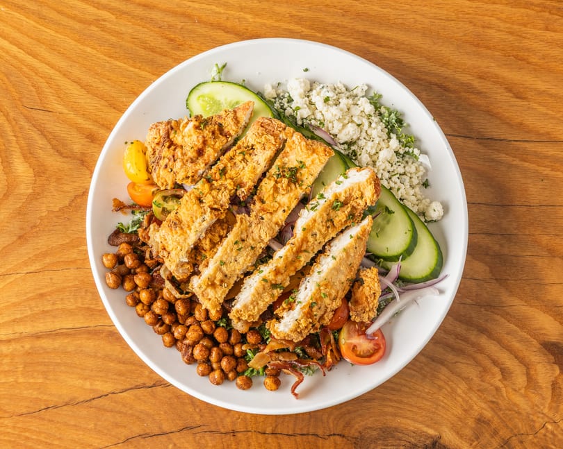 Crispy fried chicken salad with roasted chickpeas, cucumber, tomatoes, and crumbled cheese on a white plate.