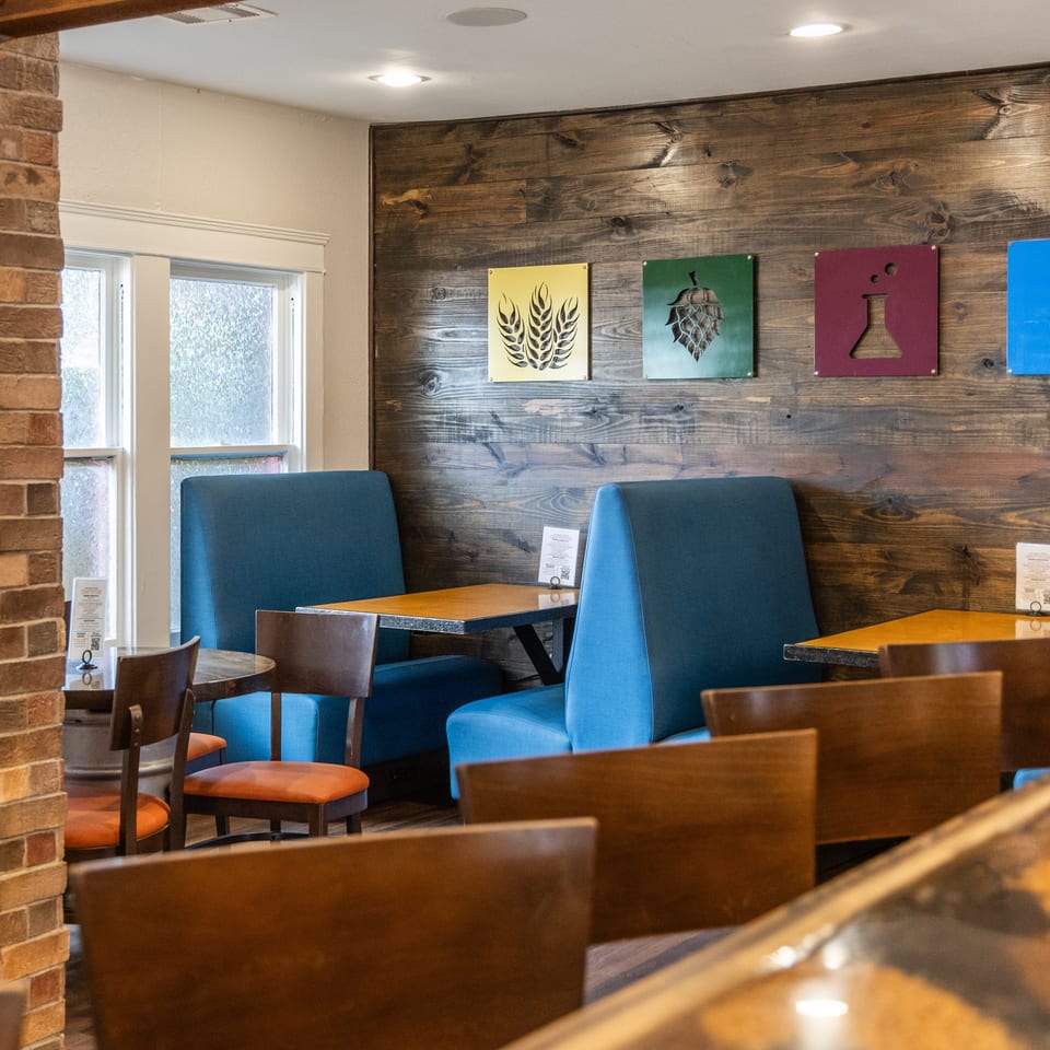 Restaurant interior with blue booths, wooden tables, and a wood-paneled wall featuring colorful beer-themed art.