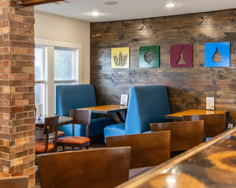 Restaurant interior with blue booths, wooden tables, and a wood-paneled wall featuring colorful beer-themed art.
