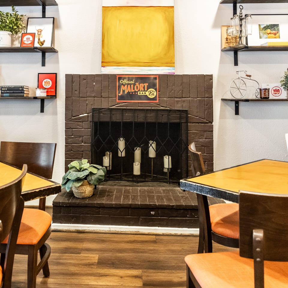 Cozy Crafthouse interior with tables, orange chairs, a brick fireplace, and "Malört Bar" sign.