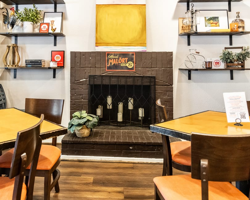 Cozy Crafthouse interior with tables, orange chairs, a brick fireplace, and "Malört Bar" sign.