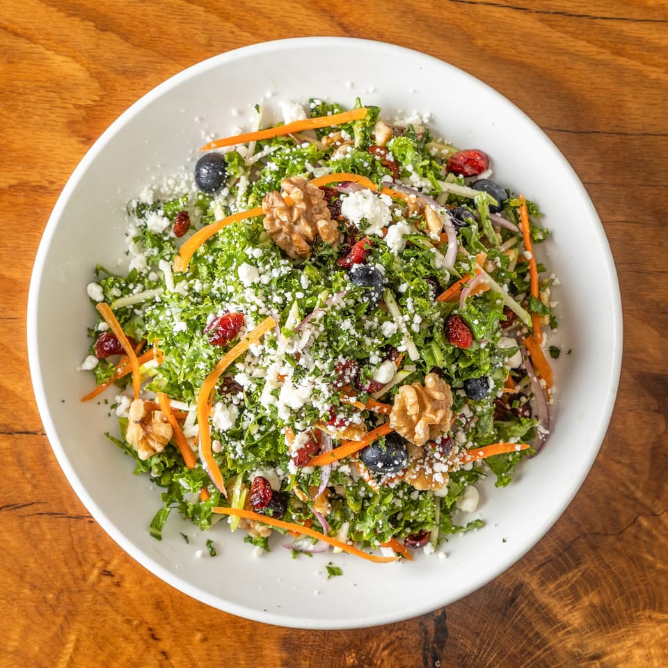 Kale salad with carrots, blueberries, cranberries, walnuts, and feta cheese in a white bowl.