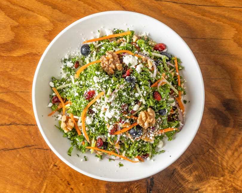 Kale salad with carrots, blueberries, cranberries, walnuts, and feta cheese in a white bowl.