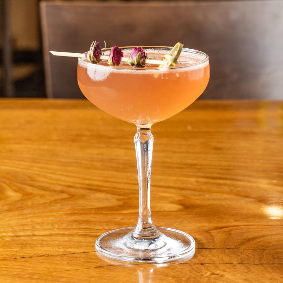 Pink cocktail in a coupe glass, garnished with dried rosebuds on a wooden table.