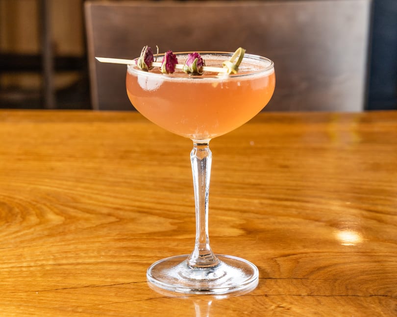 Pink cocktail in a coupe glass, garnished with dried rosebuds on a wooden table.