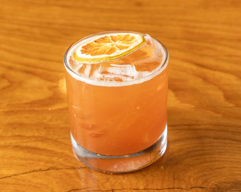 Orange cocktail with ice and a dried orange slice in a glass on a wooden table.