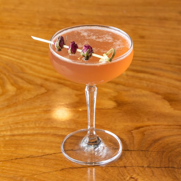 Foamy pink cocktail in a coupe glass, garnished with dried rosebuds, on a wooden table.