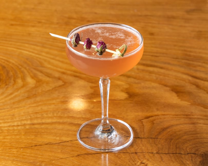 Foamy pink cocktail in a coupe glass, garnished with dried rosebuds, on a wooden table.