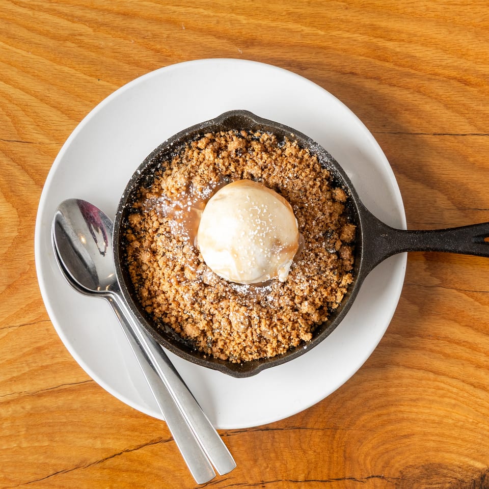 Skillet dessert with golden crumble, melting vanilla ice cream, and caramel drizzle on a wooden table.
