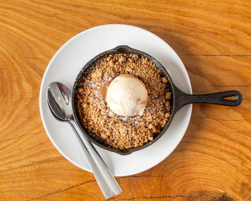 Skillet dessert with golden crumble, melting vanilla ice cream, and caramel drizzle on a wooden table.