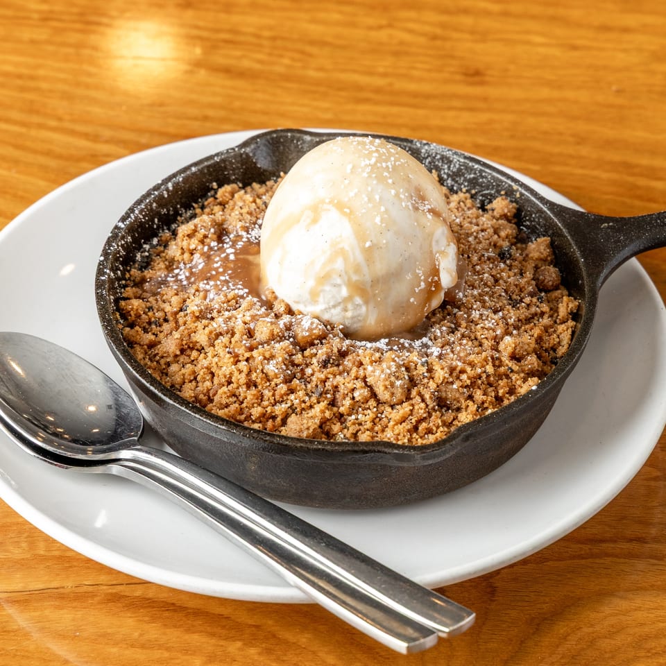 Dessert crumble in a cast iron skillet with vanilla ice cream, caramel, and powdered sugar on a wooden table.