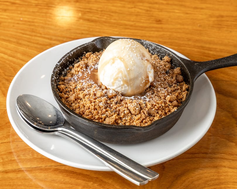Dessert crumble in a cast iron skillet with vanilla ice cream, caramel, and powdered sugar on a wooden table.