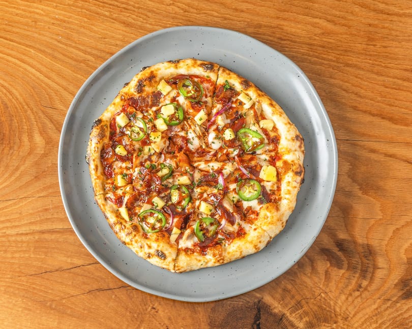 BBQ chicken pizza with pineapple, jalapeños, red onion, and bacon on a grey plate.
