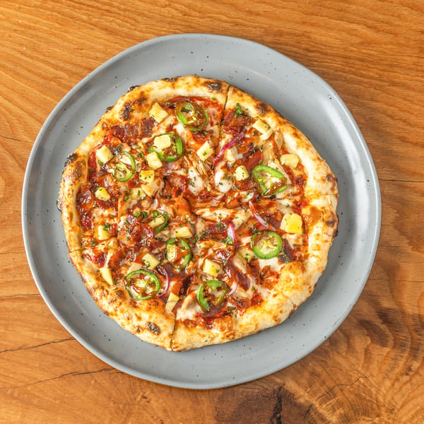 Top-down view of a BBQ chicken pizza with pineapple, jalapeños, and red onion on a grey plate.