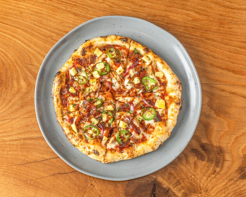 Top-down view of a BBQ chicken pizza with pineapple, jalapeños, and red onion on a grey plate.