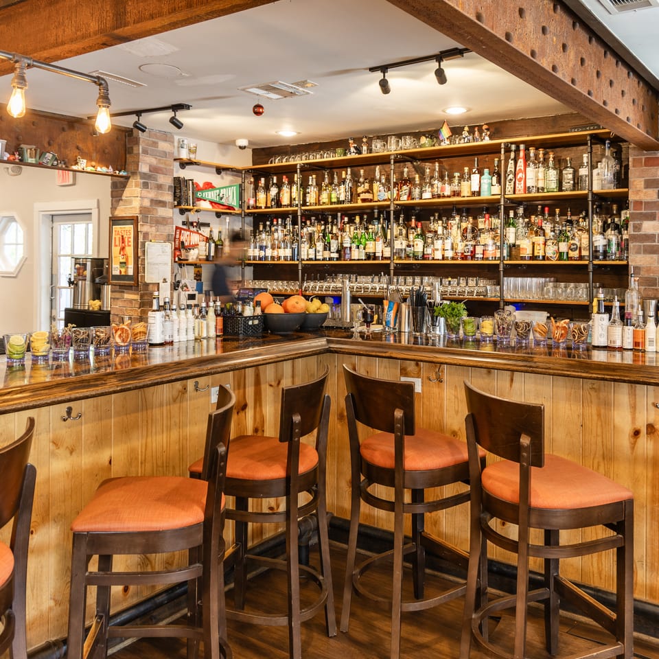 Well-stocked bar with wooden counter, orange-cushioned stools, brick accents, and a wide selection of liquor bottles.