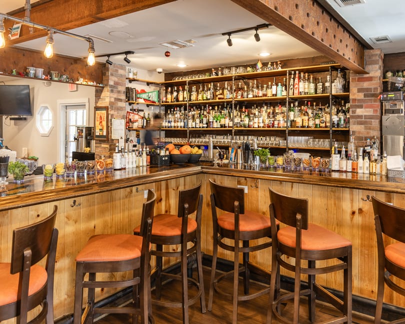 Well-stocked bar with wooden counter, orange-cushioned stools, brick accents, and a wide selection of liquor bottles.