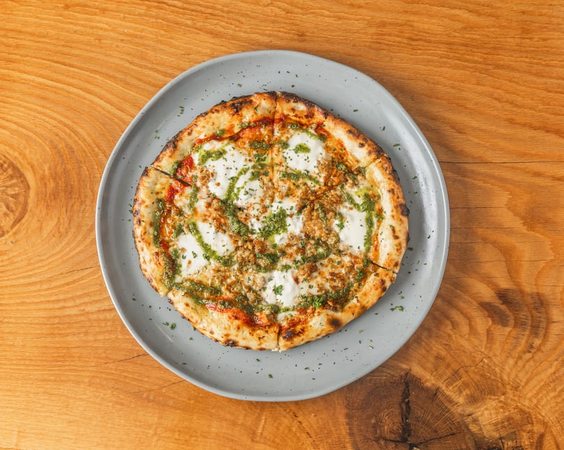Round pizza with mozzarella, pesto drizzle, and crumbled sausage on a grey plate.