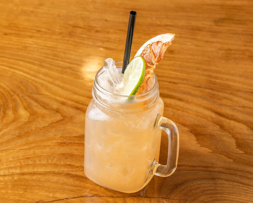 Light orange drink in a mason jar with ice, black straw, lime, and dried citrus on a wooden table.
