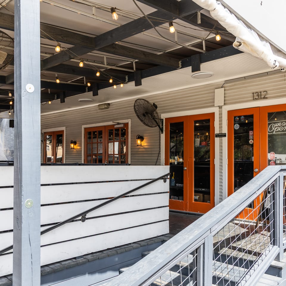 Outdoor patio of a restaurant with orange doors, 'Open' sign, string lights, and fans.