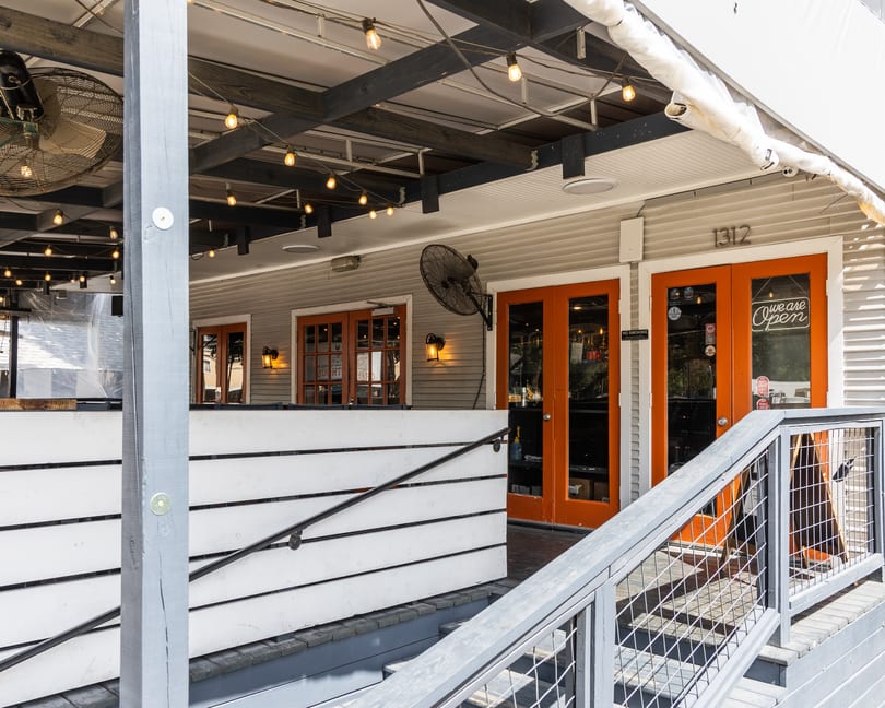 Outdoor patio of a restaurant with orange doors, 'Open' sign, string lights, and fans.