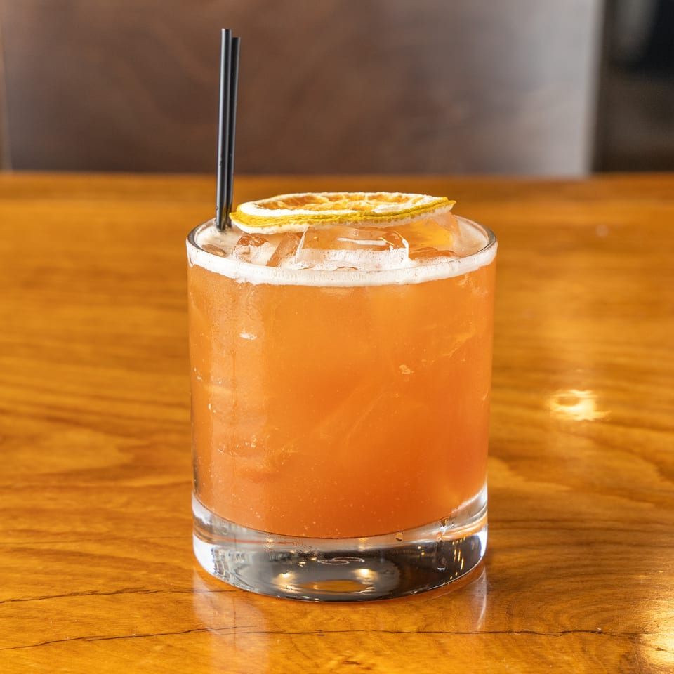 Amber cocktail with a dried orange slice and black straws on a polished wooden table.