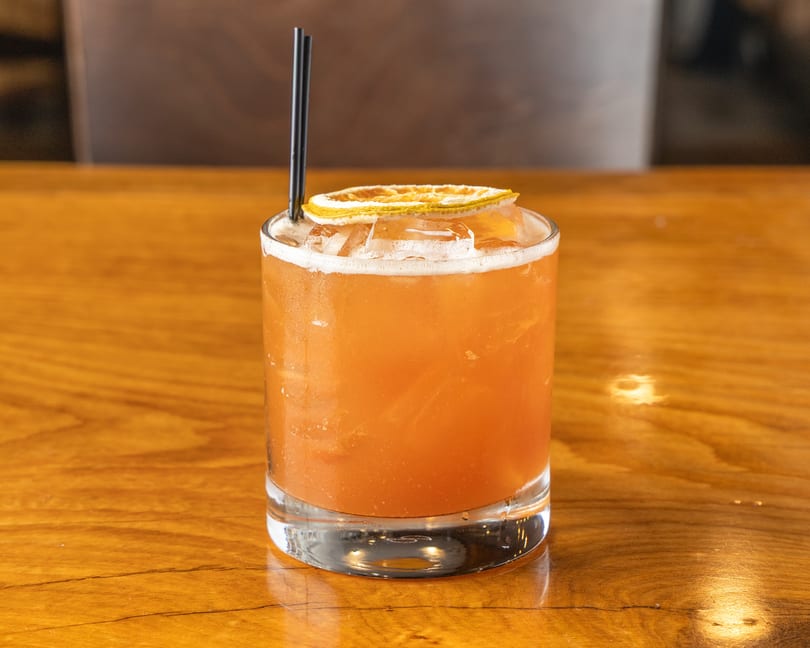 Amber cocktail with a dried orange slice and black straws on a polished wooden table.