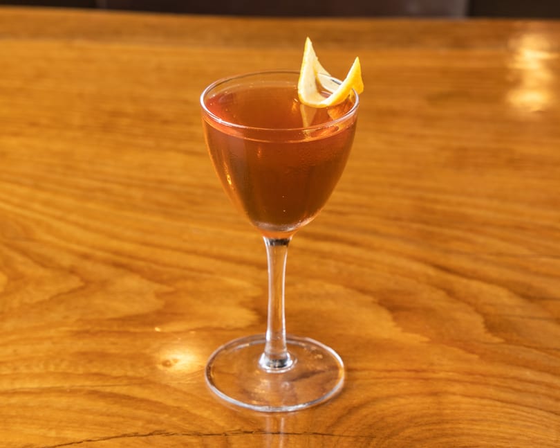 Brown cocktail in a stemmed glass, garnished with a lemon peel, on a wooden table.