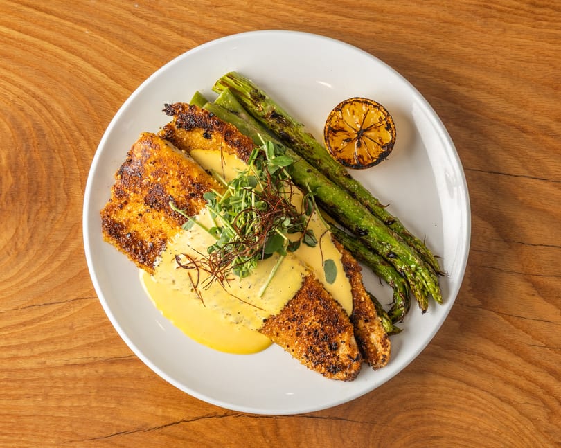 Pan-seared fish fillets with yellow sauce, microgreens, grilled asparagus, and charred lemon on a white plate.