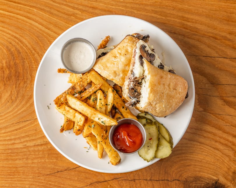 Pressed mushroom and cheese sandwich, cut open, with fries, ketchup, ranch, and pickles on a white plate.