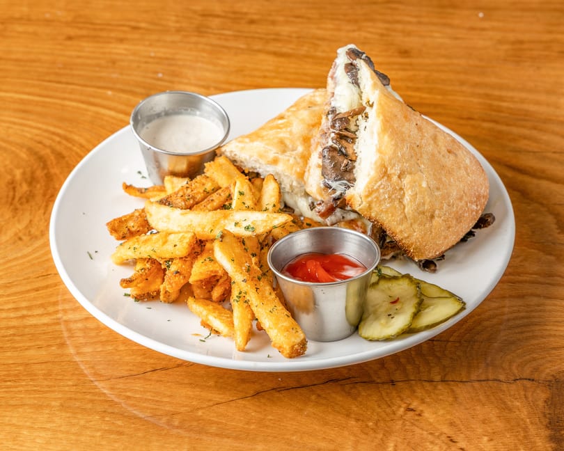 Mushroom and cheese sandwich, seasoned fries, ketchup, ranch, and pickles on a white plate.