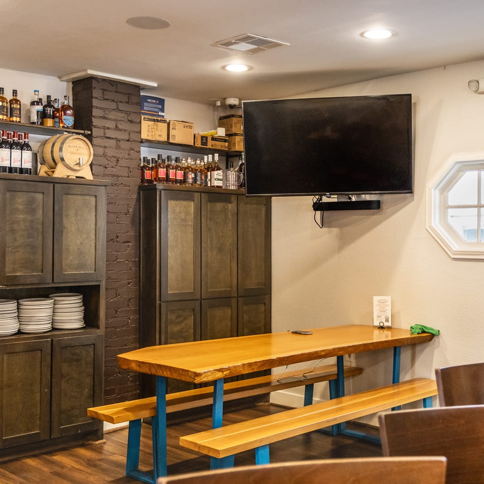 Warm restaurant interior with a large wooden communal table, benches, and dark storage cabinets.