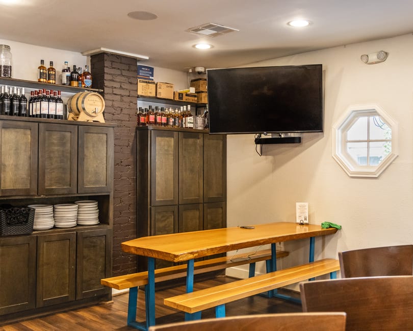 Warm restaurant interior with a large wooden communal table, benches, and dark storage cabinets.