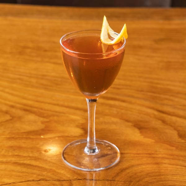 A brown cocktail in a coupe glass garnished with a lemon peel on a wooden table.