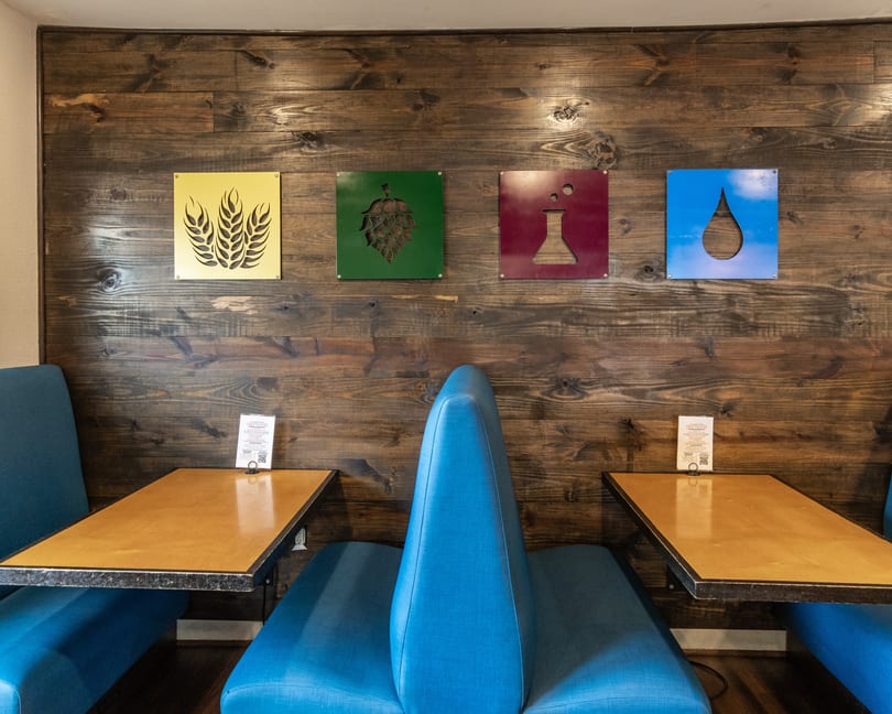 Blue booths and wooden tables in a restaurant with brewing ingredient art on a wooden wall.
